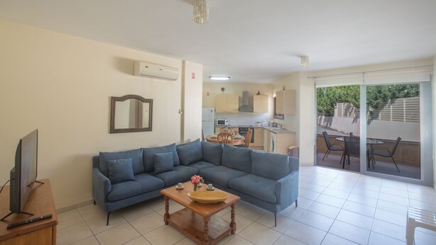 A living room with a sofa, a coffee table, and a television. There is a kitchen in the background with a dining area and a balcony with chairs. - Apt. Suri, Modern 2BDR Apt. in the heart of Ayia Napa, Close to Nissi Avenue (Ayia Napa)