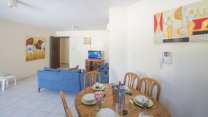 A dining area with a wooden table set for four, a television in the background, and a blue sofa in the living room. - Apt. Suri, Modern 2BDR Apt. in the heart of Ayia Napa, Close to Nissi Avenue (Ayia Napa)