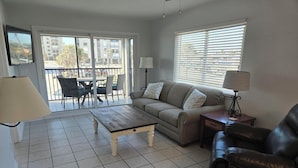 A living room with a sofa, a coffee table, a chair, and a lamp. There is a balcony with a table and chairs outside. - Estero Sands Condo with 2 bedrooms and 2 baths (Fort Myers Beach)