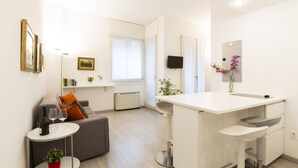 Un salon moderne avec un canapé gris, une petite table ronde sur laquelle se trouvent une bouteille de vin et des verres, et un espace cuisine blanc doté d’un comptoir-bar.