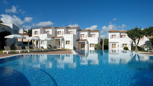 A swimming pool with lounge chairs and umbrellas, surrounded by white buildings with red-tiled roofs. - 3 beed-room Apartment (Manacor)