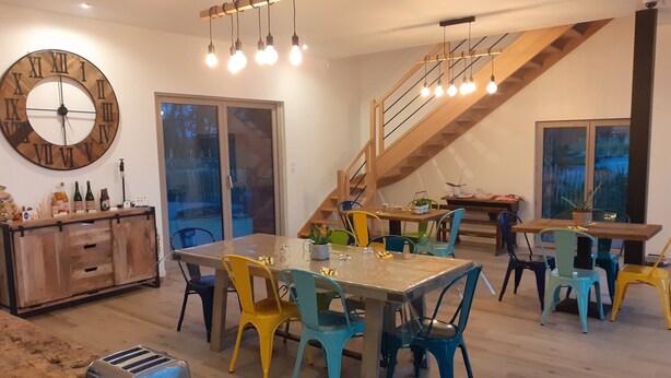 Un comedor amplio con una mesa de madera y sillas coloridas, un reloj de pared grande y una escalera que sube a un nivel superior.