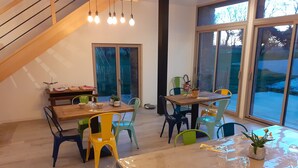 Un comedor con mesas de madera y sillas coloridas, ventanas amplias y una escalera.