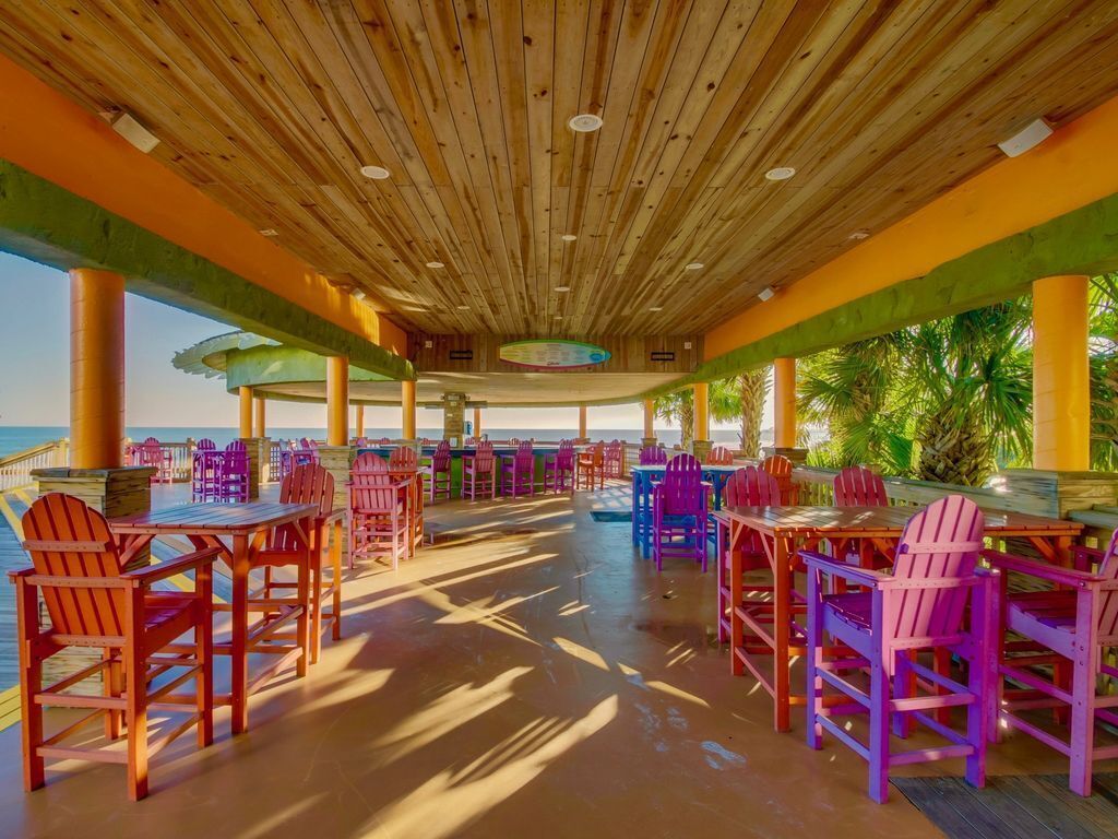 An outdoor dining area with wooden tables and chairs, surrounded by a wooden ceiling and columns, overlooking a beach.