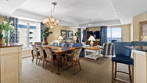A spacious dining area with a wooden table and chairs, a chandelier, and a view of the city through large windows. - Royale Palms 1201 2 Br Oceanfront in Kingston,Pool (Myrtle Beach)