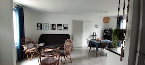 Een woonkamer met een bank, twee rieten stoelen, een eettafel met stoelen, en een keuken met een koelkast en een hangplant.