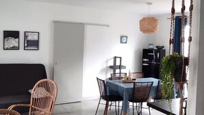A modern living room with a dining area, featuring wicker furniture, a hanging light fixture, and a kitchen in the background. - Vacation home 3* LES MATHES/LA PALMYRE (Les Mathes)