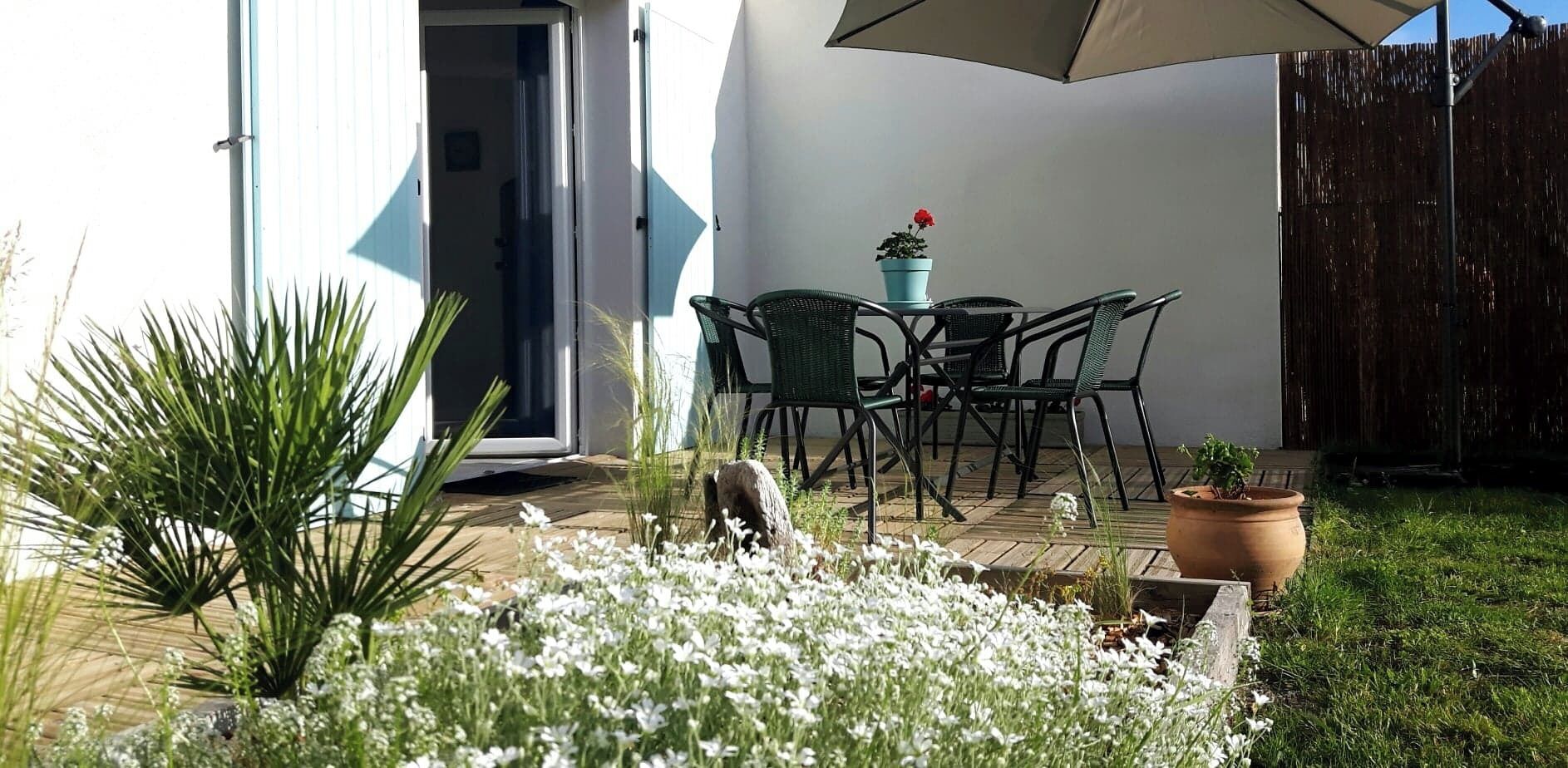 Pátio externo com mesa e cadeiras, rodeado por plantas em vasos e cerca de madeira.