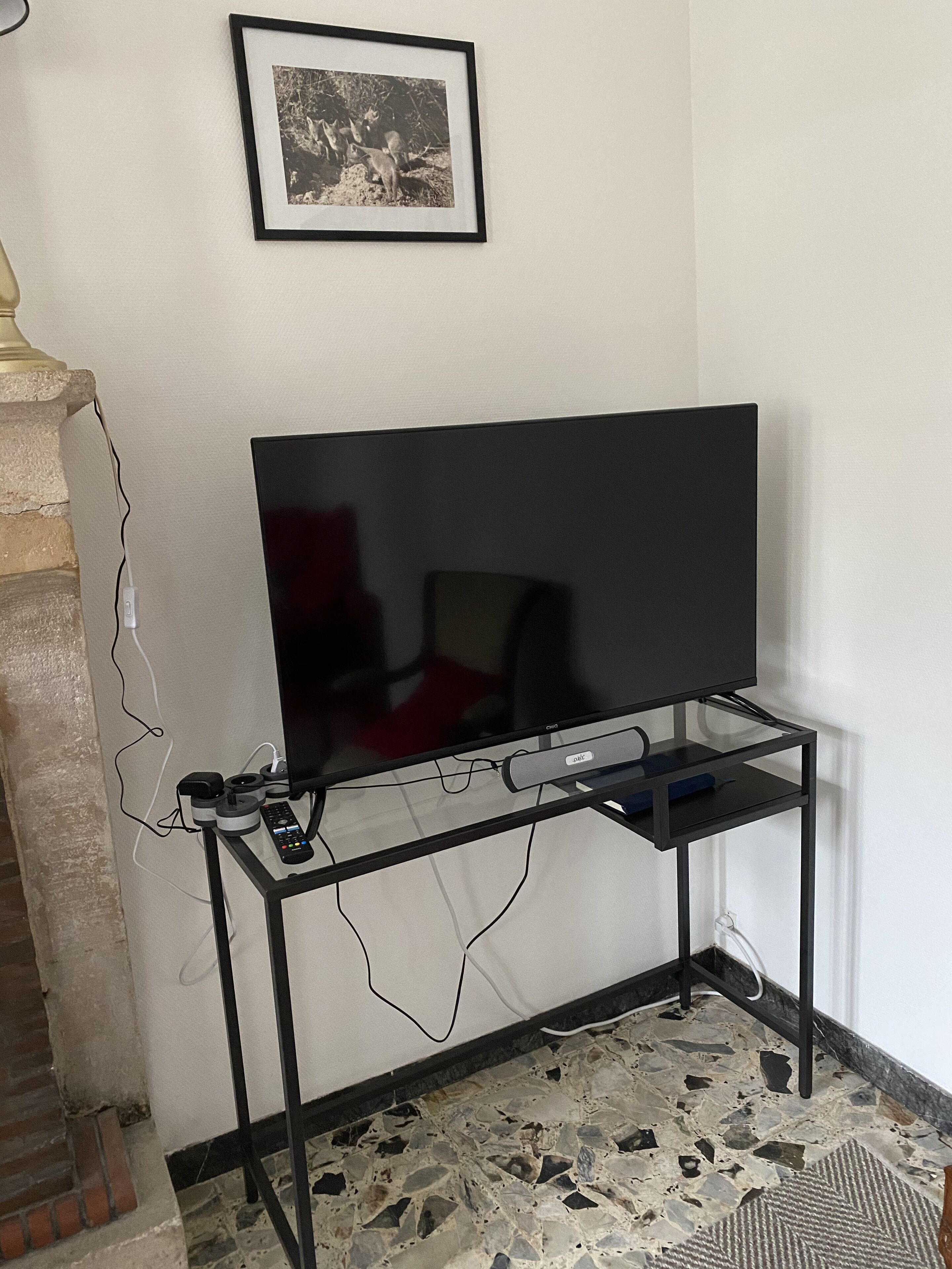 A television on a black metal stand with a remote and cables, a framed picture on the wall, and a stone-tiled floor.