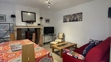 A living room with a red sofa, a coffee table, a TV, and a fireplace. There is a dining table with chairs and a patterned tablecloth.