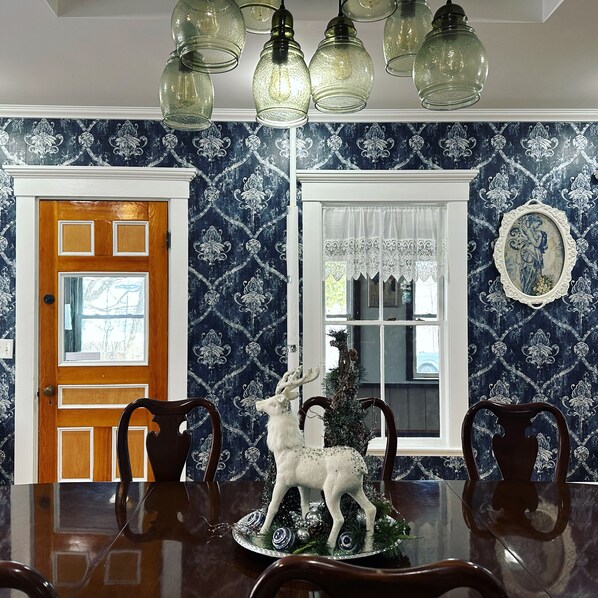 A dining room with a wooden table, chairs, and a decorative deer centerpiece. There are two windows with curtains, a door, and a wall with a patterned wallpaper. - Spacious Historic Home in the White Mountains, great for Groups, Sleeps 14 (Bartlett)