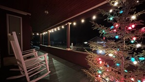 A covered porch with a rocking chair, a Christmas tree with lights, and a view of a house in the background.