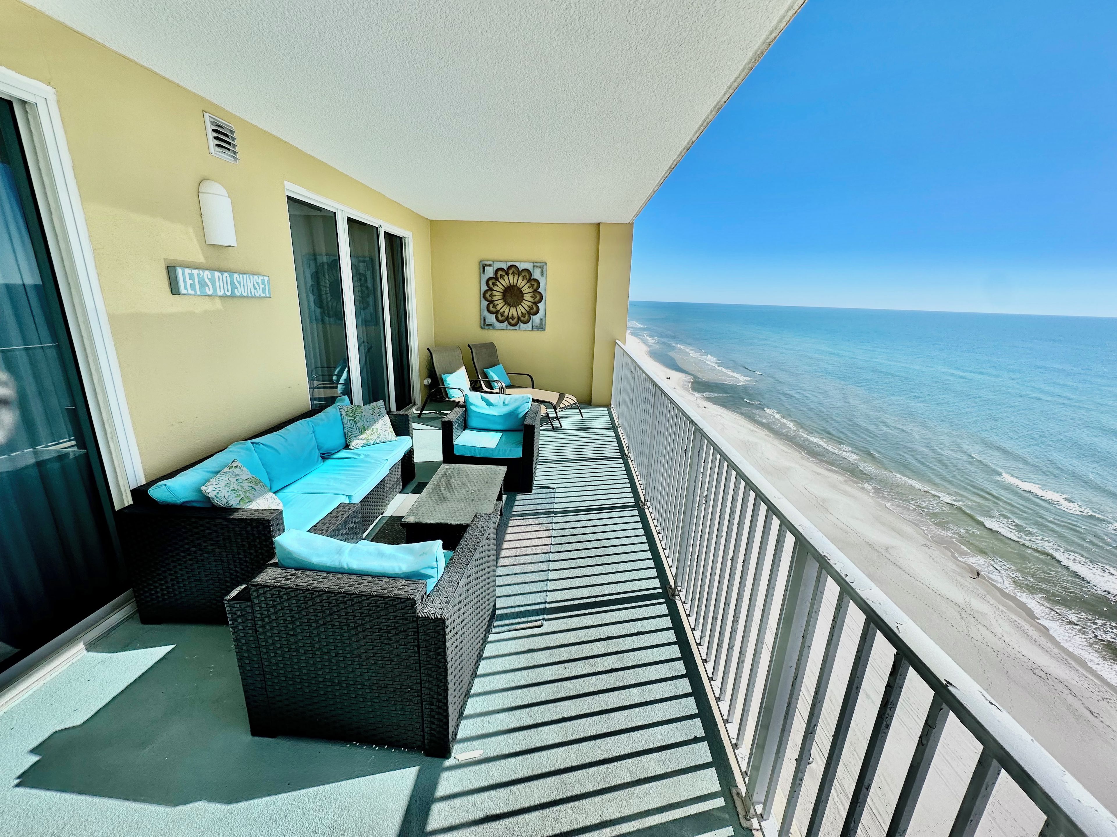 Un balcone con mobili in vimini, vista sulla spiaggia e un cartello con su scritto 'LET'S DO SUNSET'.