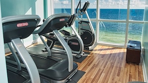 Uma academia com esteiras e equipamentos de exercícios, janelas amplas com vista para o oceano.