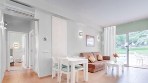 Ein modernes Wohnzimmer mit einem weißen Esstisch und Stühlen, einem Sofa und einem Couchtisch. Eine Glastür führt auf einen Balkon mit Blick ins Grüne.