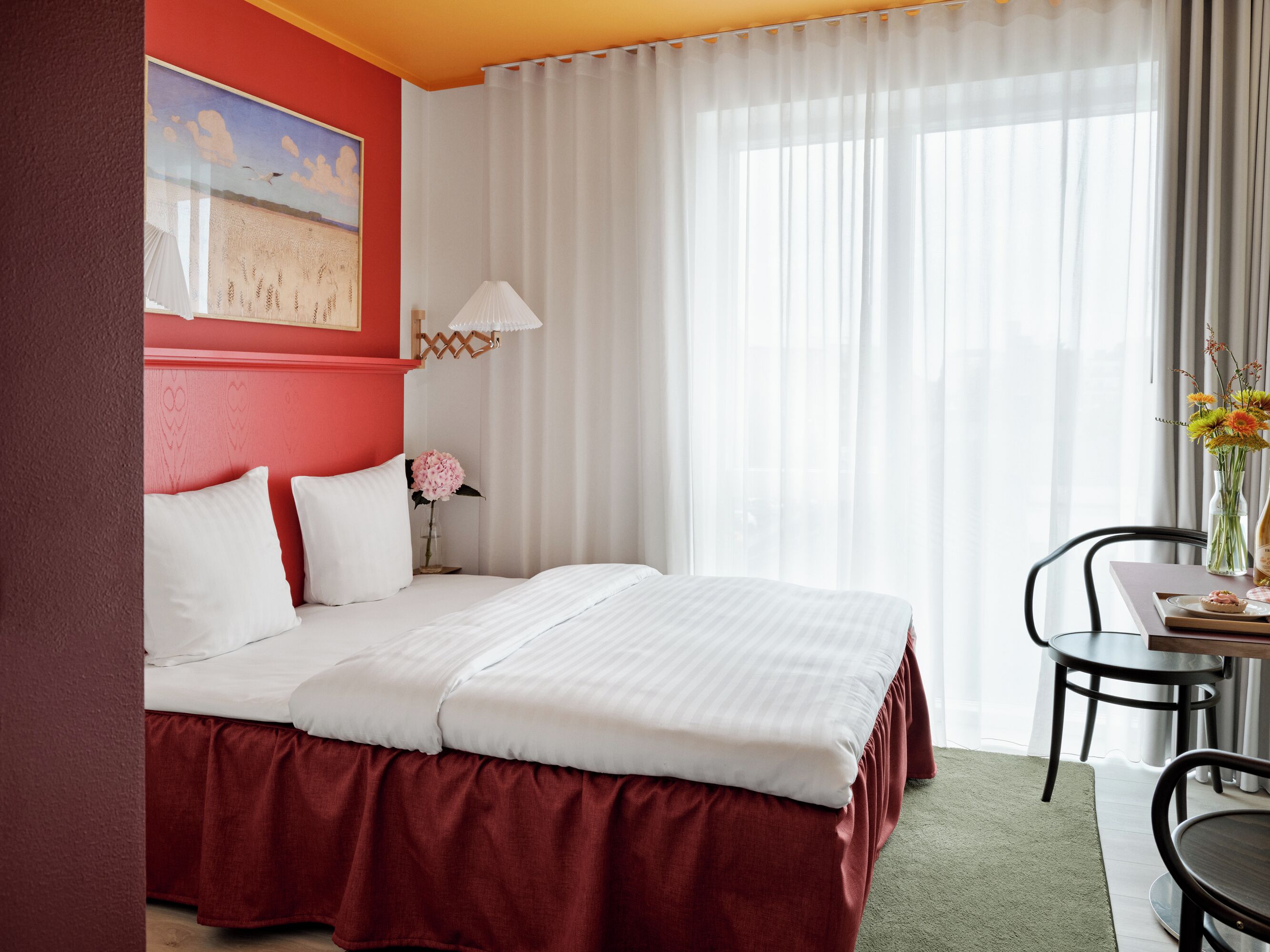A hotel room with a large bed, a desk with a chair, a vase with flowers, and a painting on the wall.