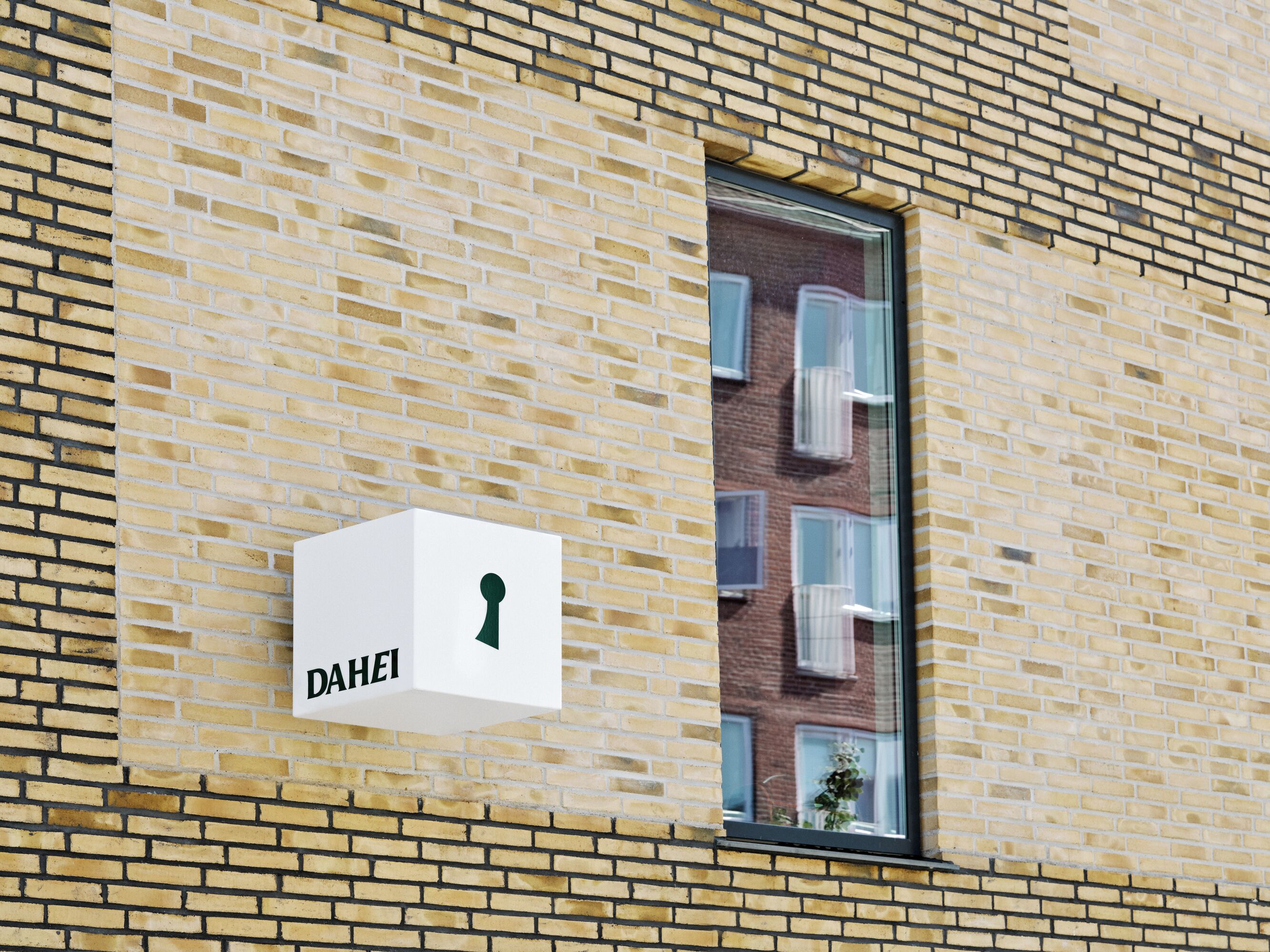 A white sign with the word "DAHEI" on a brick wall, next to a window with white blinds.