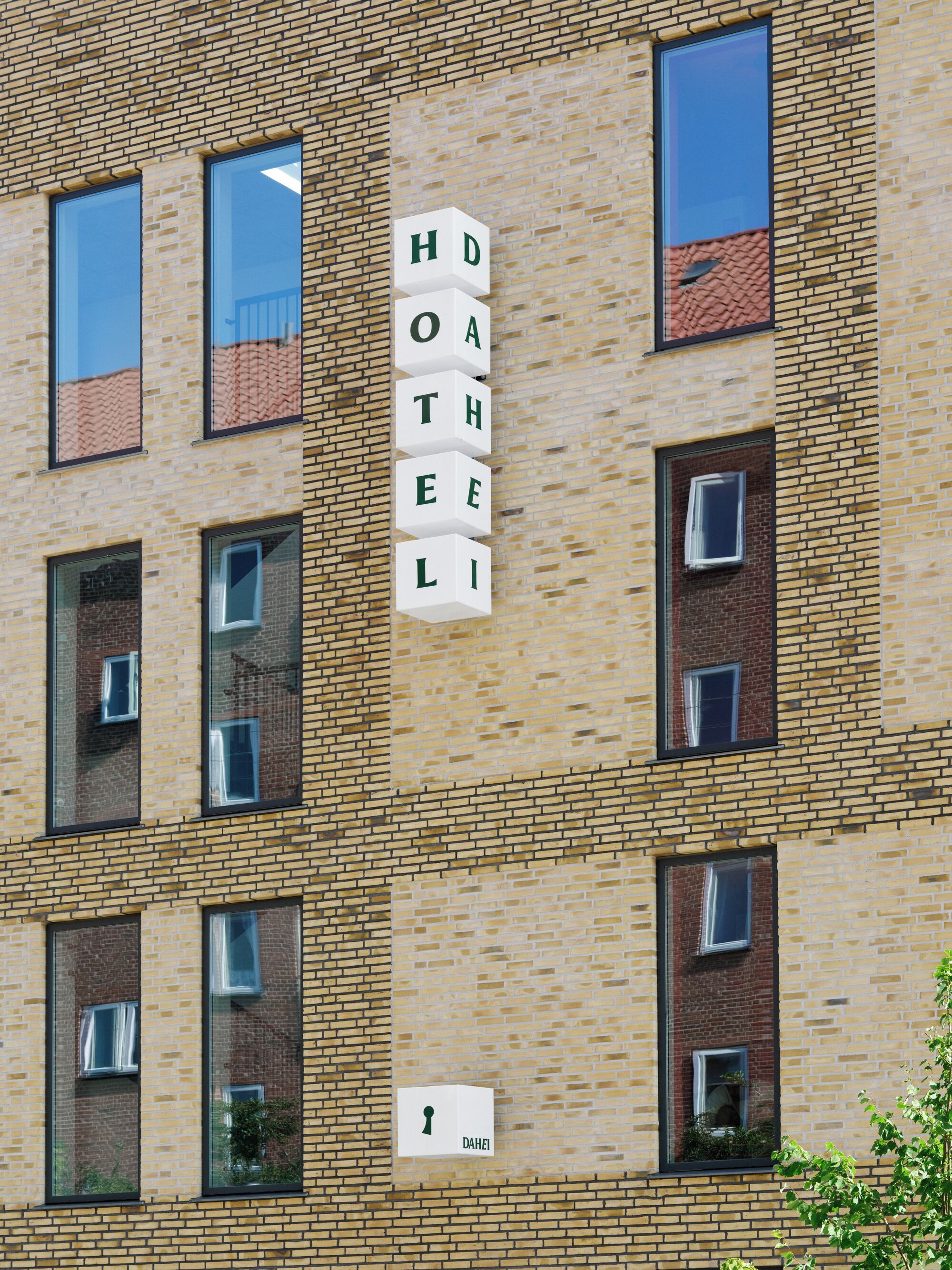 En byggnad med flera våningar, en fasad av tegel och stora fönster. På byggnaden finns en skylt med bokstäverna ”H O A T E L I”, vilket visar att det är ett hotell. Under hotelskylten finns en symbol som föreställer ett nyckelhål.