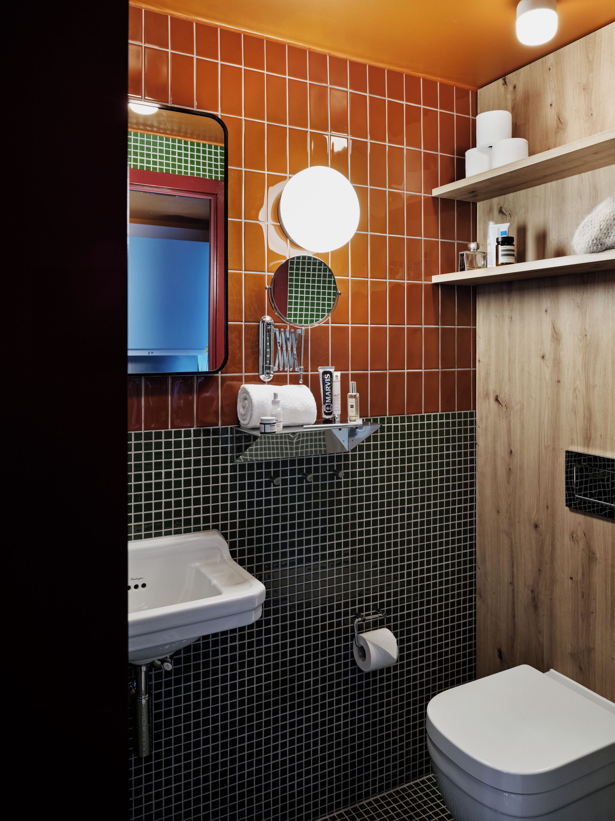 Et badeværelse med en hvid vask, et spejl, et toilet og en hylde med toiletartikler.