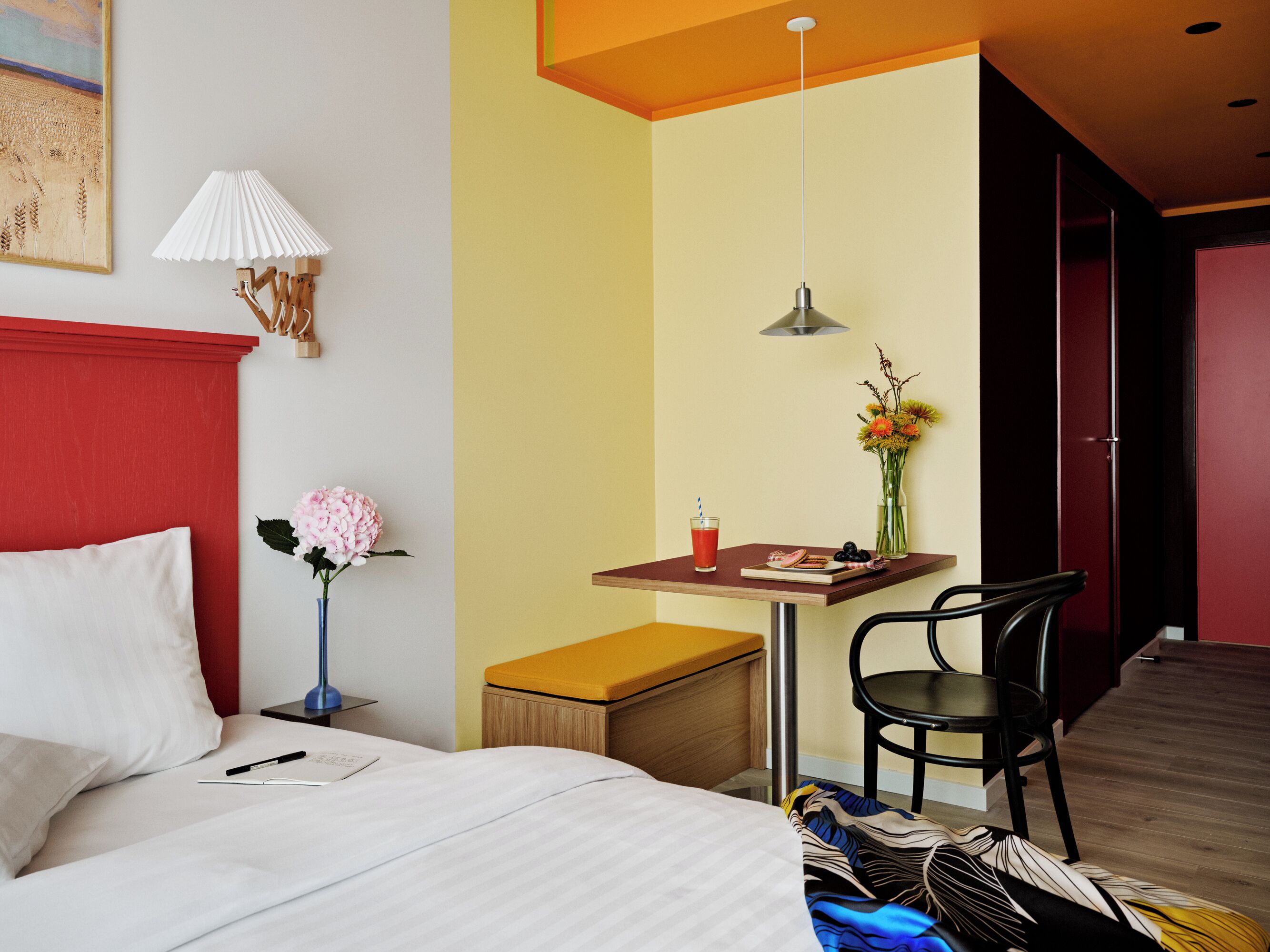 Una habitación de hotel con una cama, una mesita con bebidas y aperitivos, una silla, una lámpara y un jarrón con flores.