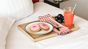 Una cama bien hecha con una almohada blanca, una bandeja con galletas y un vaso de jugo, un florero con flores y una pared roja.