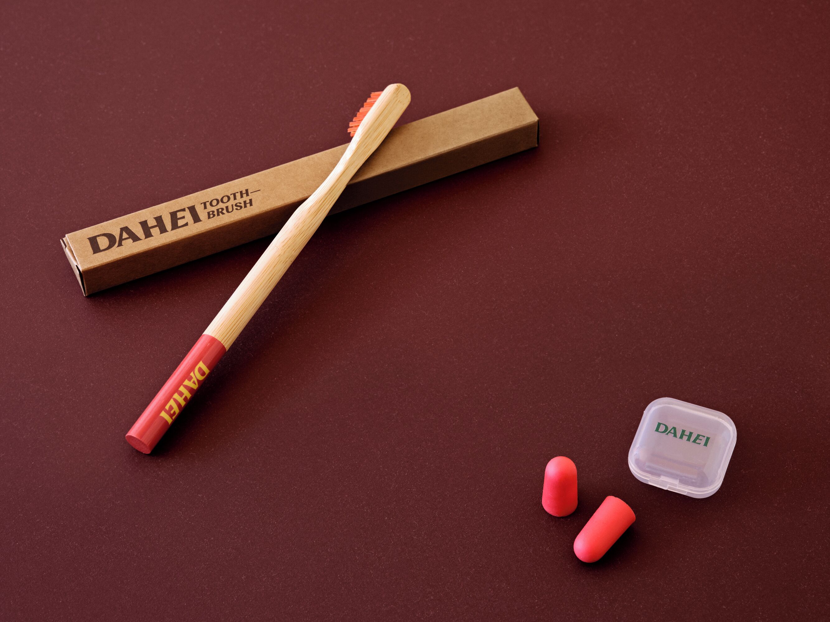 Un cepillo de dientes DAHEI, un estuche para cepillo de dientes y dos cápsulas rojas sobre una superficie color borgoña.