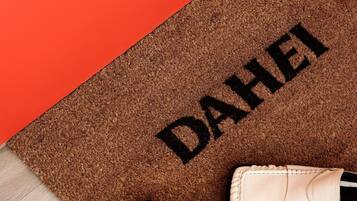 A brown doormat with "DAHEI" printed on it, placed on a wooden floor next to a pair of white shoes.
