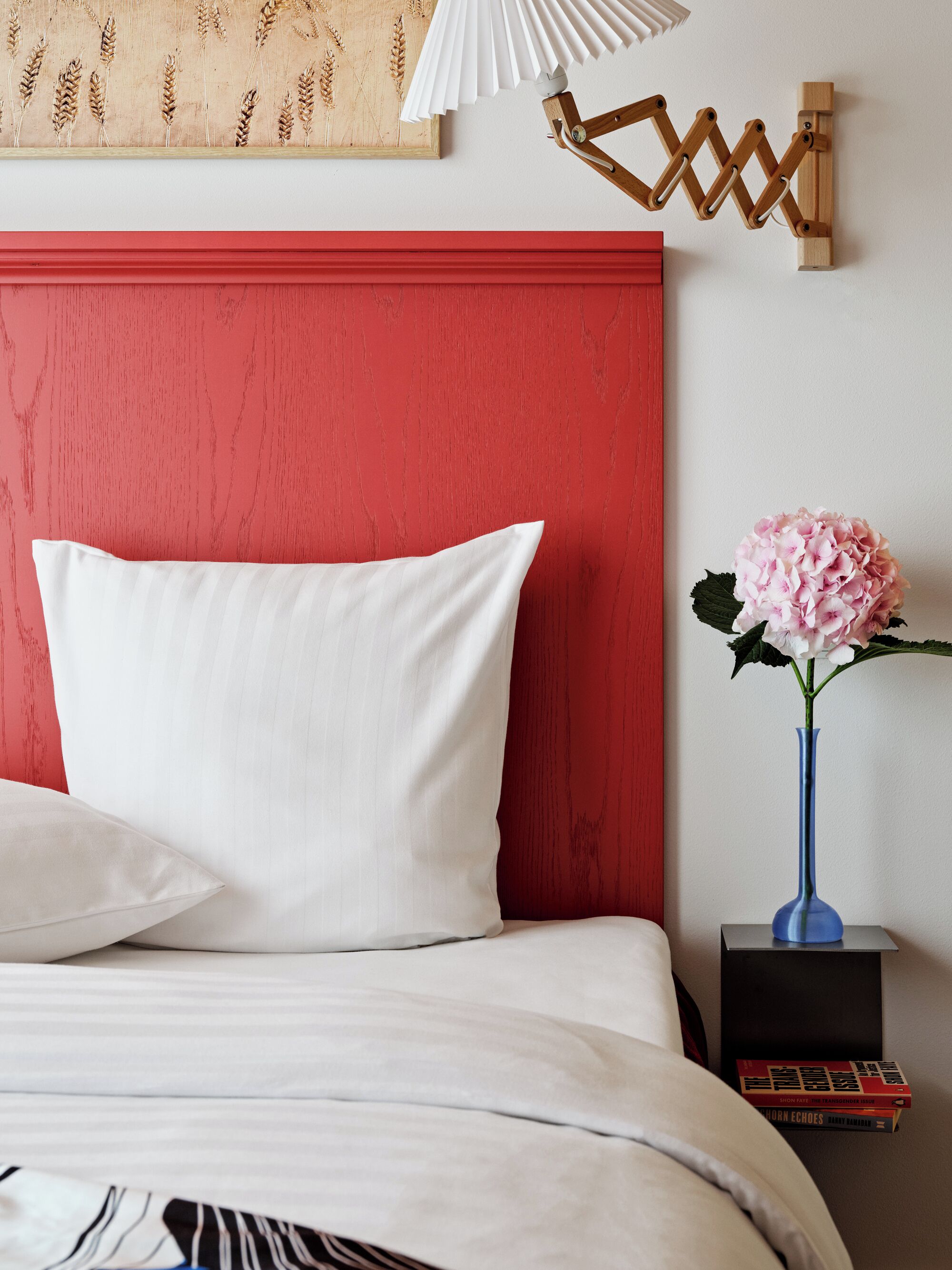 Een net opgemaakt bed met witte lakens, een rode hoofdbord en een blauwe vaas met roze bloemen op een nachtkastje.