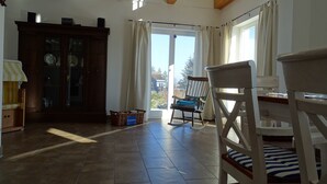 Une pièce avec un plafond en bois, une table à manger avec des chaises, un fauteuil à bascule, un meuble avec des portes en verre et un panier posé sur le sol.