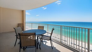A balcony with a table and chairs, overlooking a beach with clear blue water and a sandy shore.