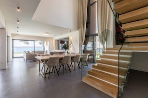 A modern interior with a dining area, staircase, and large windows overlooking a body of water. - Luxury Seafront Villa Papillon Split with Pool (Kaštel Kambelovac)