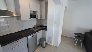Een compacte keuken met een magnetron, spoelbak en vaatwasser. Er staat een prullenbak en een klein zithoekje met twee stoelen.