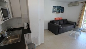 Een compacte leefruimte met een kitchenette, een grijze bank met oranje kussens en een klein tafeltje.