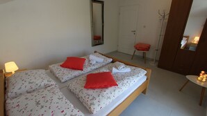 Una stanza con due letti singoli, ognuno con un cuscino rosso e un asciugamano arrotolato. Si vede uno specchio a parete, una porta e un piccolo sgabello con una federa rossa.