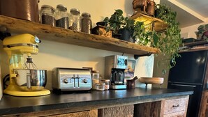 A kitchen with wooden cabinets, a black countertop, a yellow mixer, a toaster, a coffee maker, and shelves with jars and plants. - The Farmhouse At Shilo Farm, 4 miles from Portsmouth, 10 minutes from the Beach (Eliot)