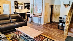 Een gezellige woonkamer met een leren bank, een houten salontafel en een vloerkleed met dierenprint. Er is een keuken met houten kastjes en een eettafel met stoelen. Een groot schilderij hangt aan de muur, en er is een decoratie van een hertenhof.