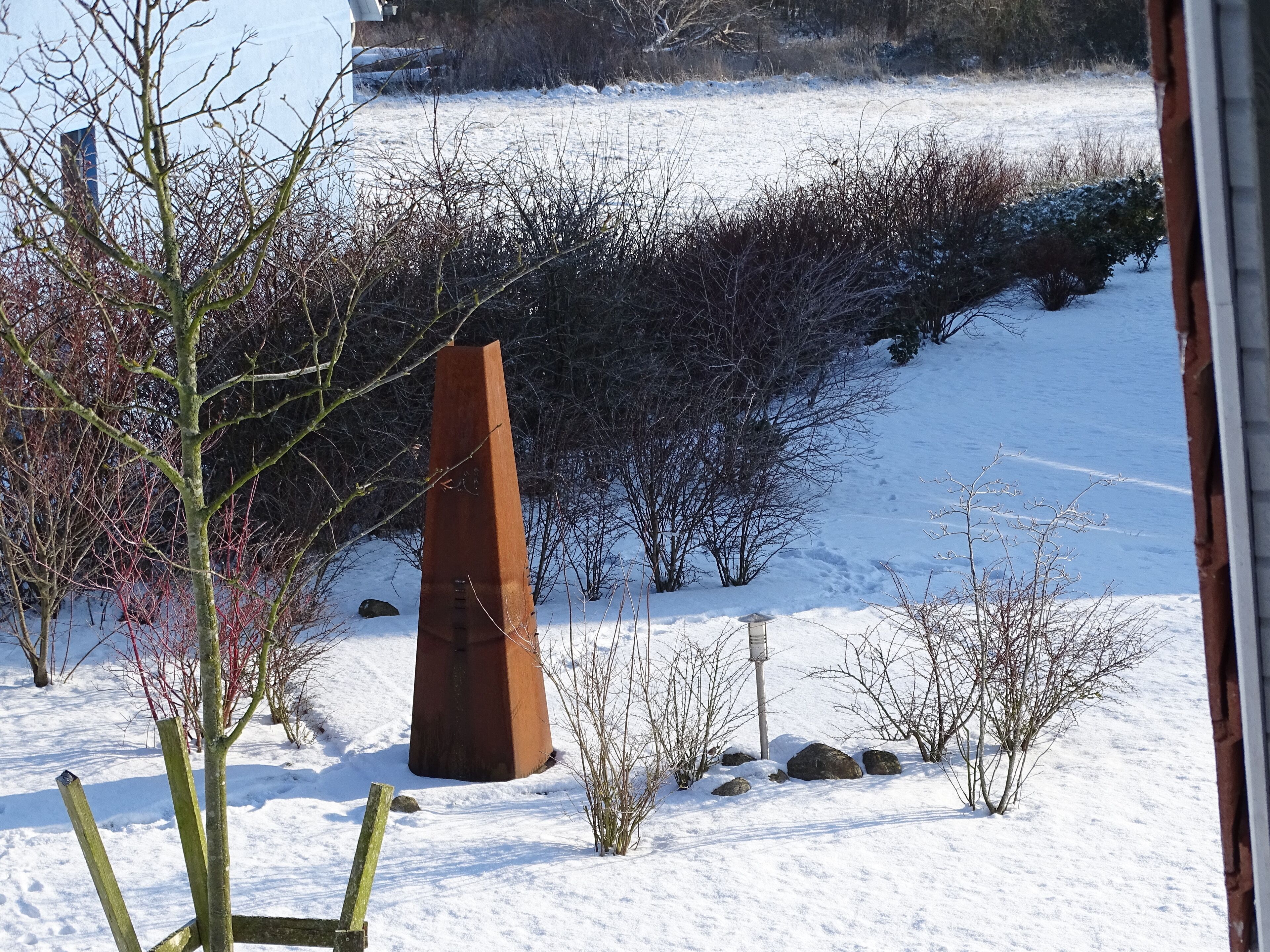 A snow-covered landscape with a tall, rusted metal sculpture in the foreground, surrounded by bare trees and shrubs.
