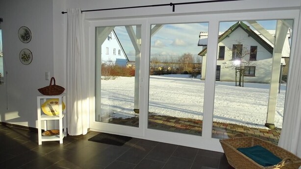 A room with a large sliding glass door leading to a snowy outdoor area, a wicker chair, and a small white shelf with a basket. - thatched house, direct sea view, idyllic location in vacation region Usedom (Freest, Usedom)