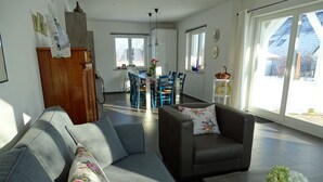 A living room with a grey sofa, a brown armchair, and a coffee table. There is a dining area in the background with a table and chairs. - thatched house, direct sea view, idyllic location in vacation region Usedom (Freest, Usedom)