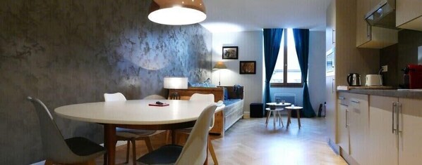 A dining area with a round table and chairs, a sofa, and a lamp. There is a kitchen area with cabinets and a window with curtains.