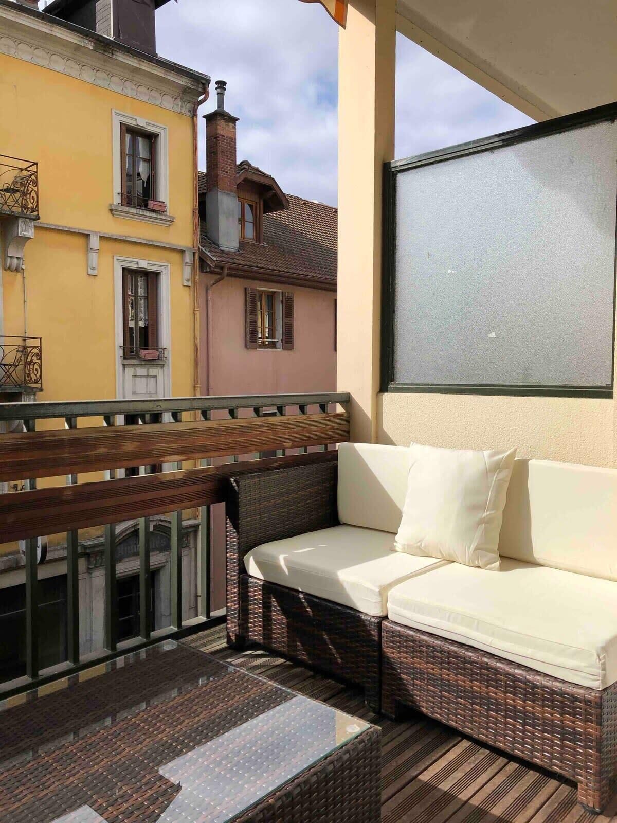 A balcony with a wicker sofa and a glass table, overlooking a yellow building with a chimney.