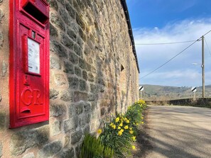 A stone building with a red post box and yellow flowers by the roadside.