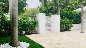 A landscaped garden with a paved walkway, a palm tree, and a white gate. - Beach Getaway, Cozy Studio Villa #2 (Lauderdale-by-the-Sea)