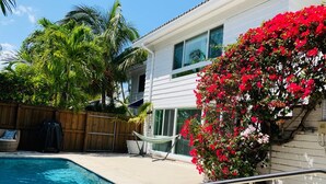 A swimming pool with a wooden fence, a hammock, and a house with a white exterior and red flowering plants. - Beach Getaway, Cozy Studio Villa #2 (Lauderdale-by-the-Sea)