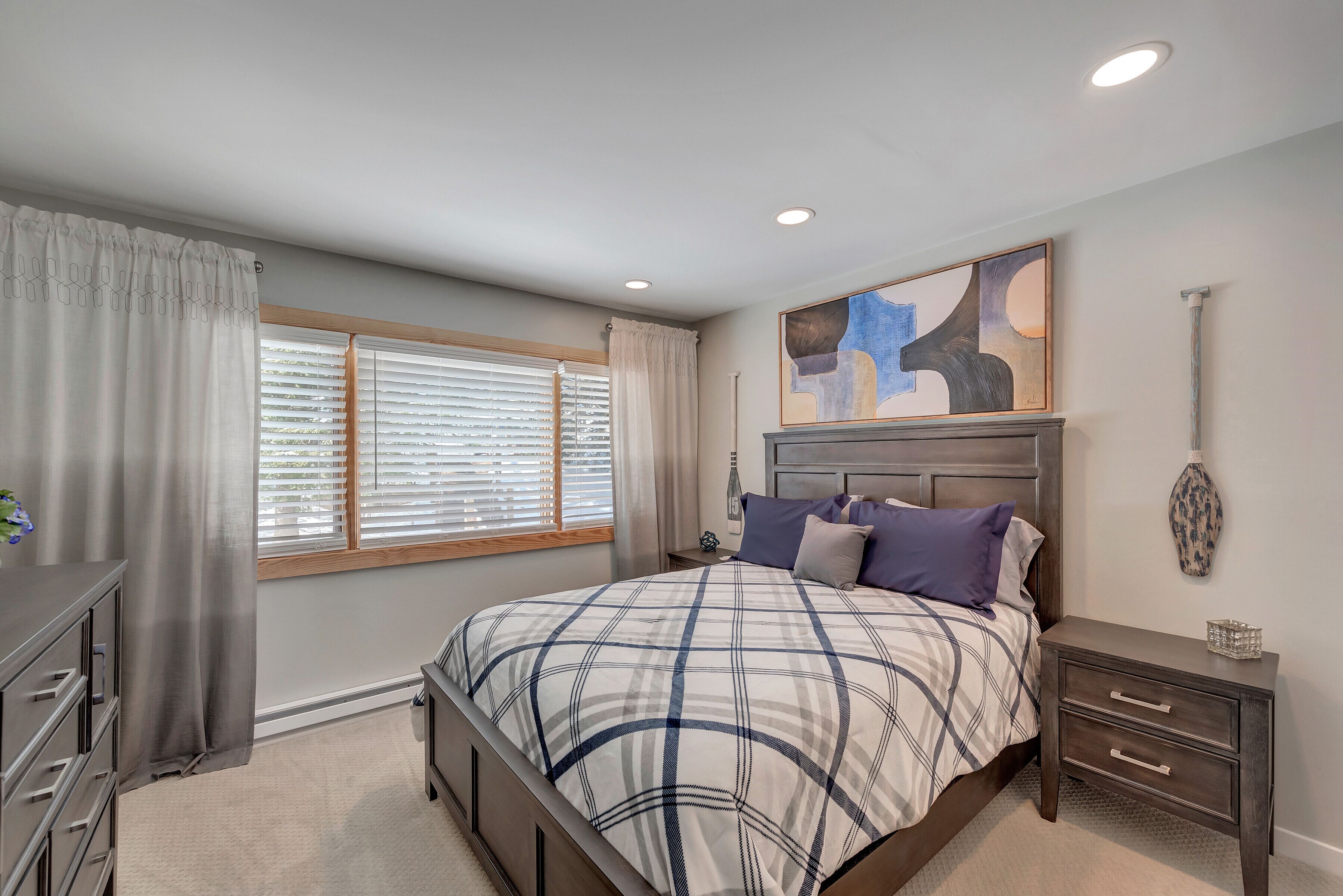 Une chambre à coucher comprenant un lit, des tables de chevet, une commode et une fenêtre avec des stores.