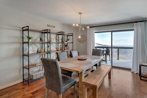 A dining area with a wooden table, chairs, and a bench. There is a bookshelf with various decorative items and a view of the outdoors through large windows.