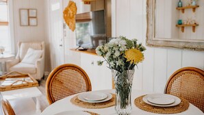 Ein Essbereich mit einem runden Tisch, gedeckt für vier Personen, umgeben von Korbstühlen. Auf dem Tisch steht eine Vase mit Blumen, im Hintergrund sind Regale mit Dekorationen zu sehen.