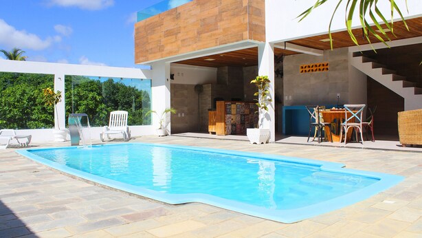 A modern outdoor pool area with a clear blue pool, lounge chairs, and a dining area with outdoor furniture. - Casa Barramares em Cotovelo com piscina (Parnamirim)