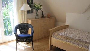 Une chambre à coucher avec un lit en bois, une chaise en osier avec un coussin, une commode, une lampe et un vase rempli de fleurs.