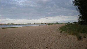 Une plage de sable fin bordée d’une étendue d’eau calme, quelques arbres épars et un bâtiment visible au loin.